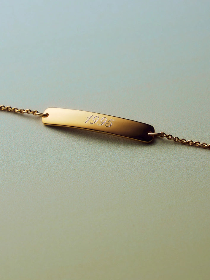 Gold bracelet with a bar charm engraved with '1995' on a light green background