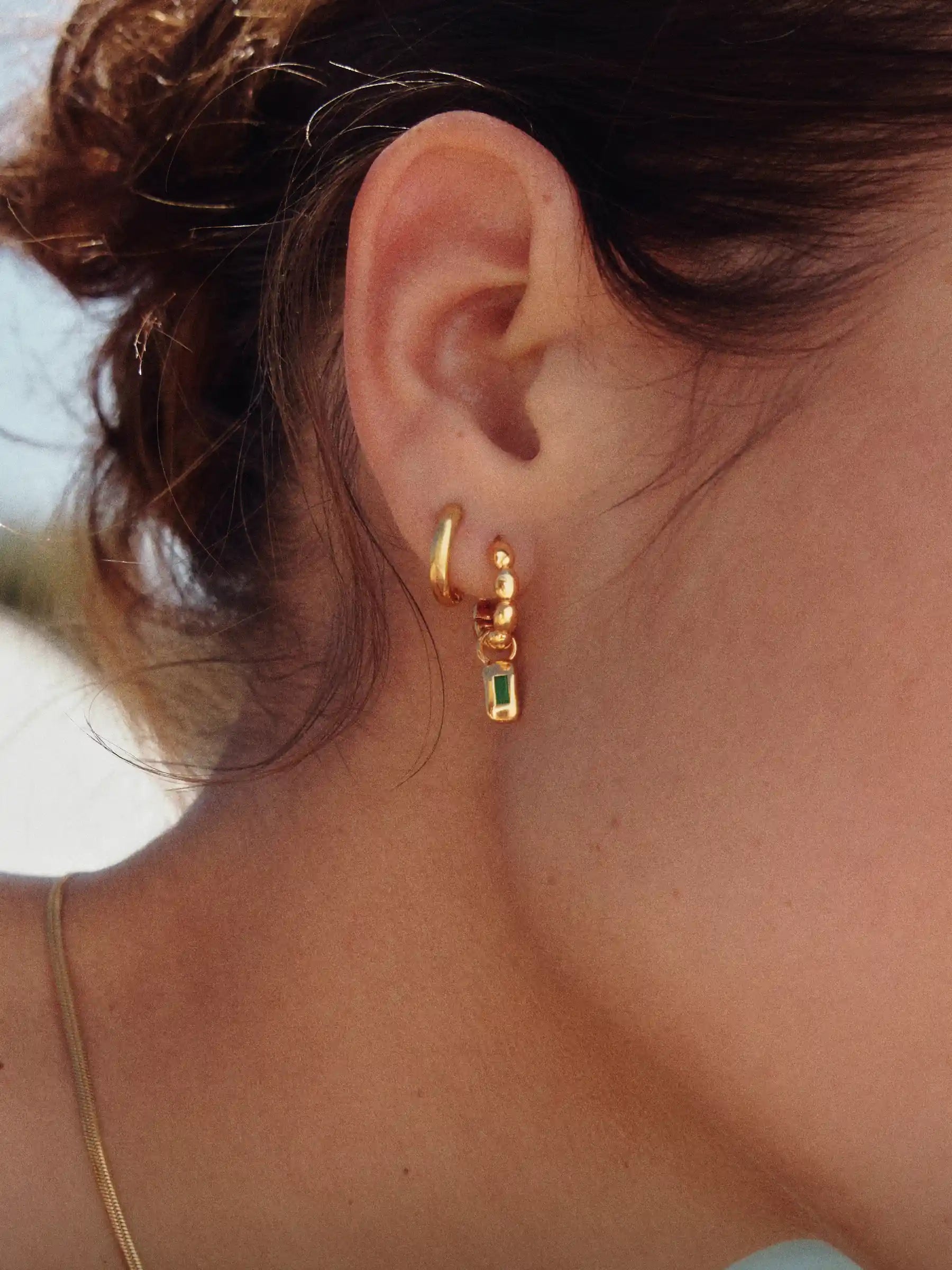 Close-up of an ear wearing gold hoop earrings with a green gemstone.