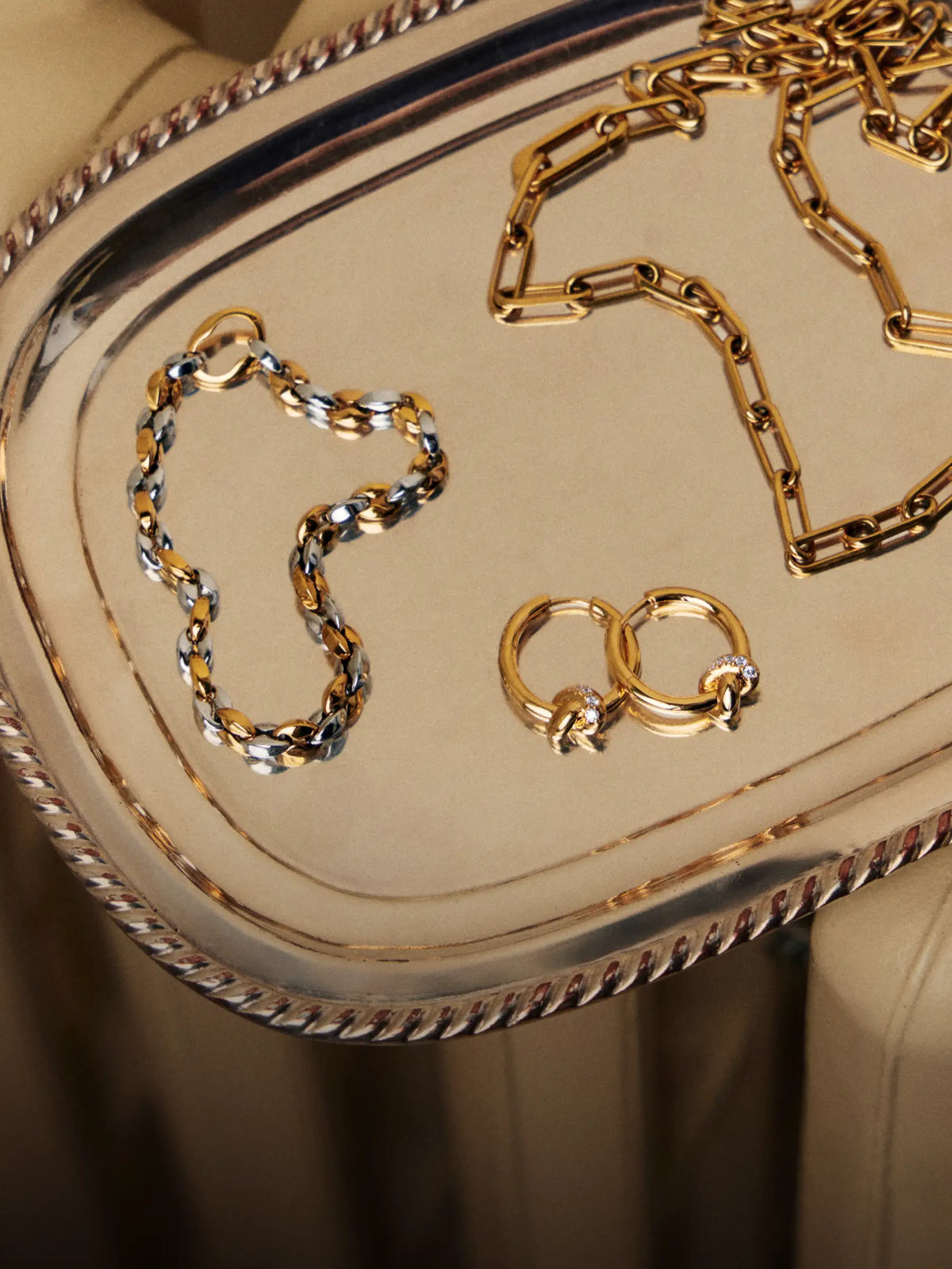 Gold and silver chain jewelry pieces, including a chain necklace, chain bracelet, and two gold rings, arranged on a reflective silver tray.